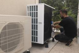 Technician servicing an outdoor air conditioner unit beside a second unit on a sunny patio.
